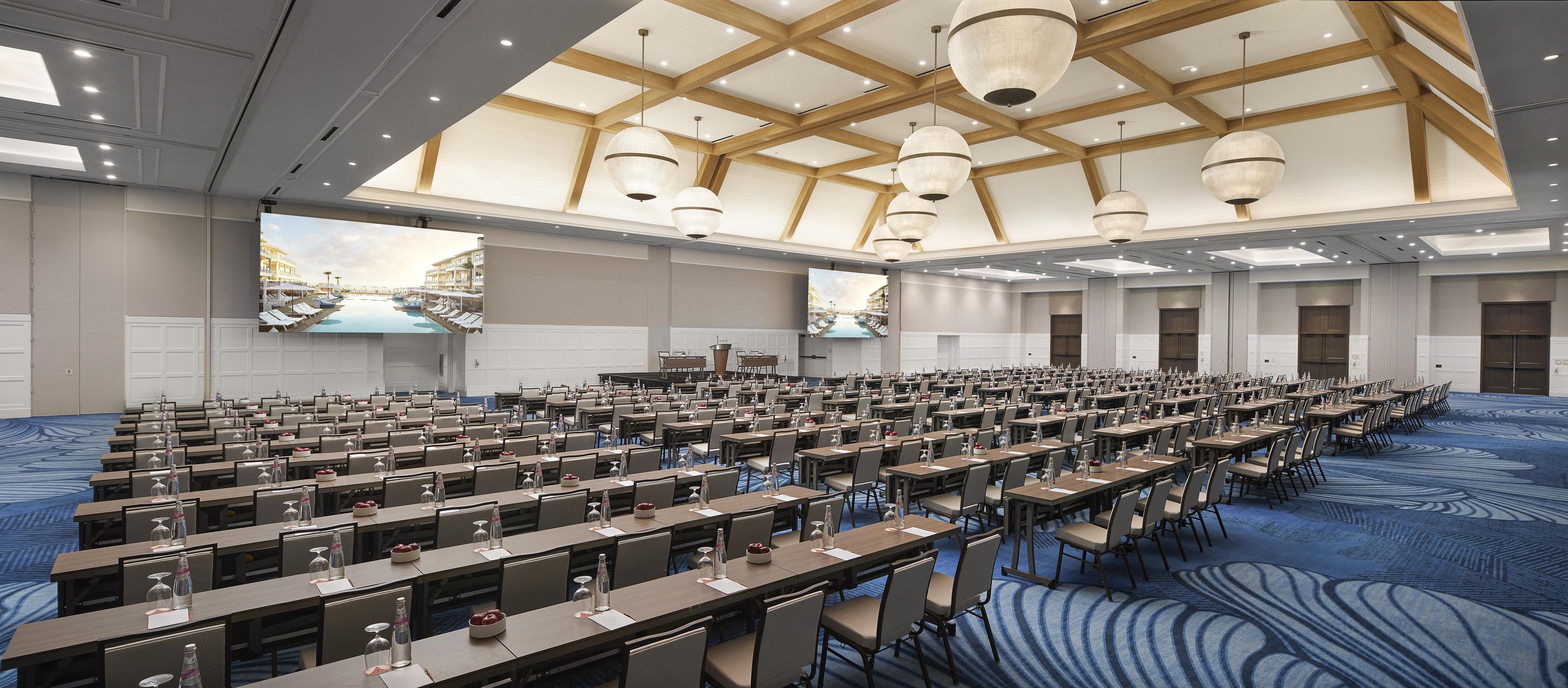A large conference room with multiple tables and chairs, a high ceiling with wooden beams, and a large screen displaying a scenic view.
