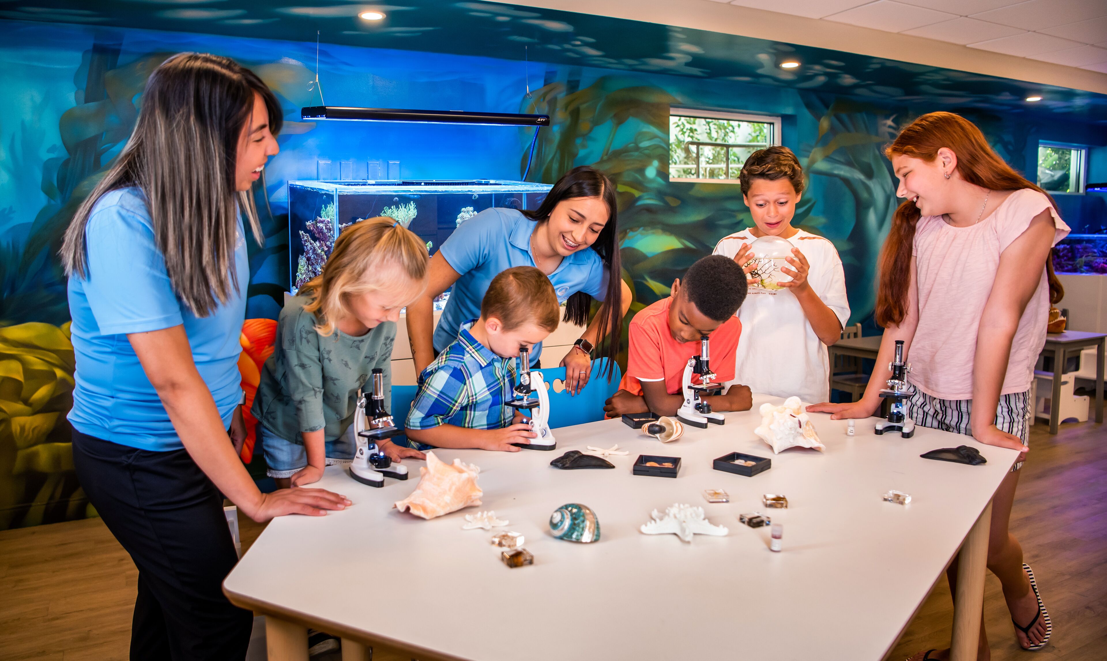 A group of people engaged in a science activity with microscopes and specimens on a table in a room with educational murals.