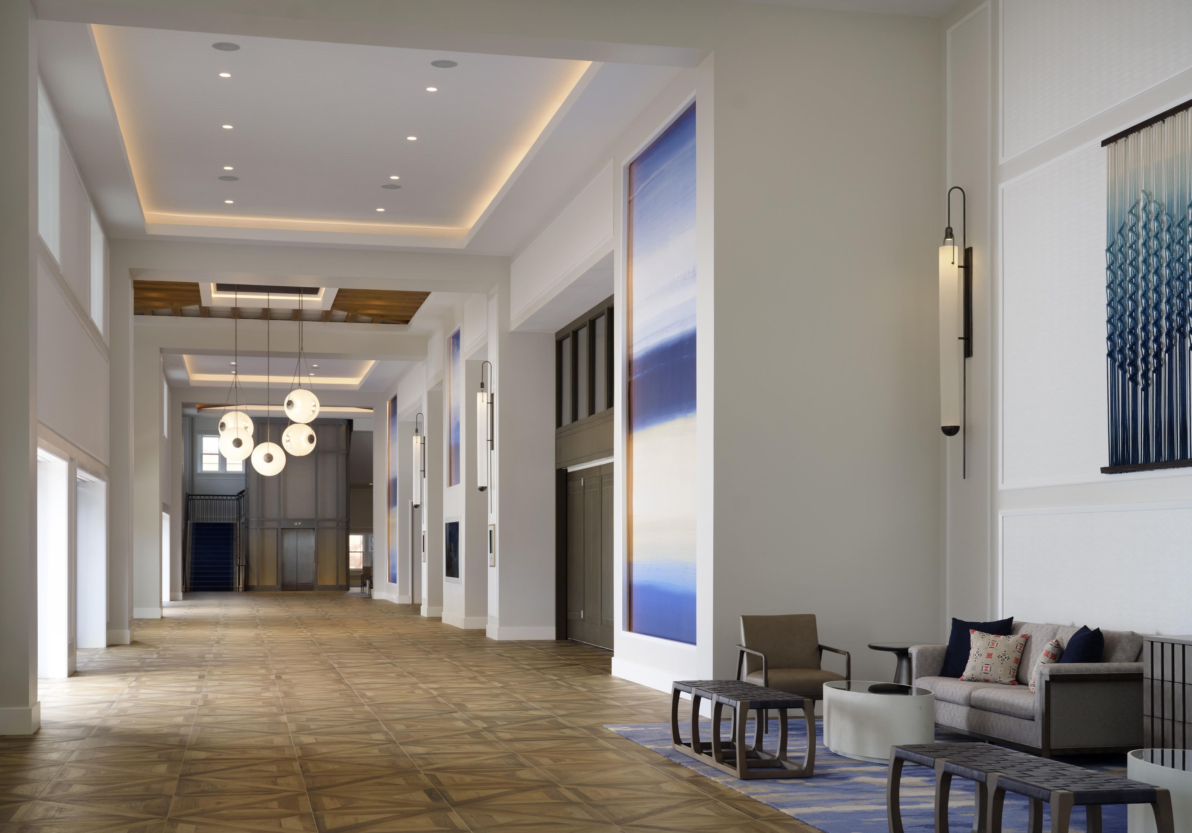 Un vestíbulo de hotel con un largo pasillo, luminarias modernas, un mostrador de recepción y una zona de estar con un sofá y una mesa de centro.