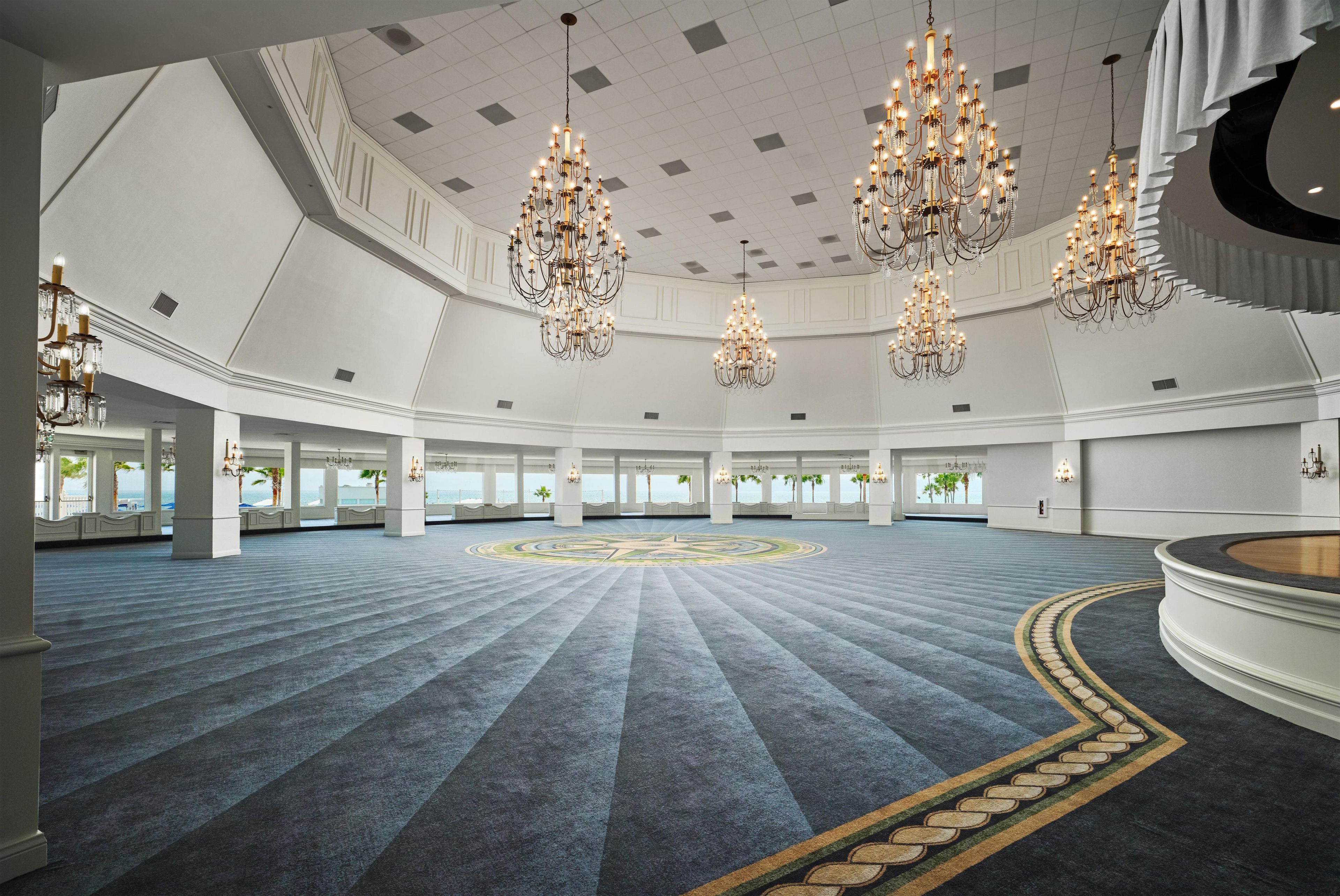 A spacious hall with a patterned carpet, multiple chandeliers, and large windows.