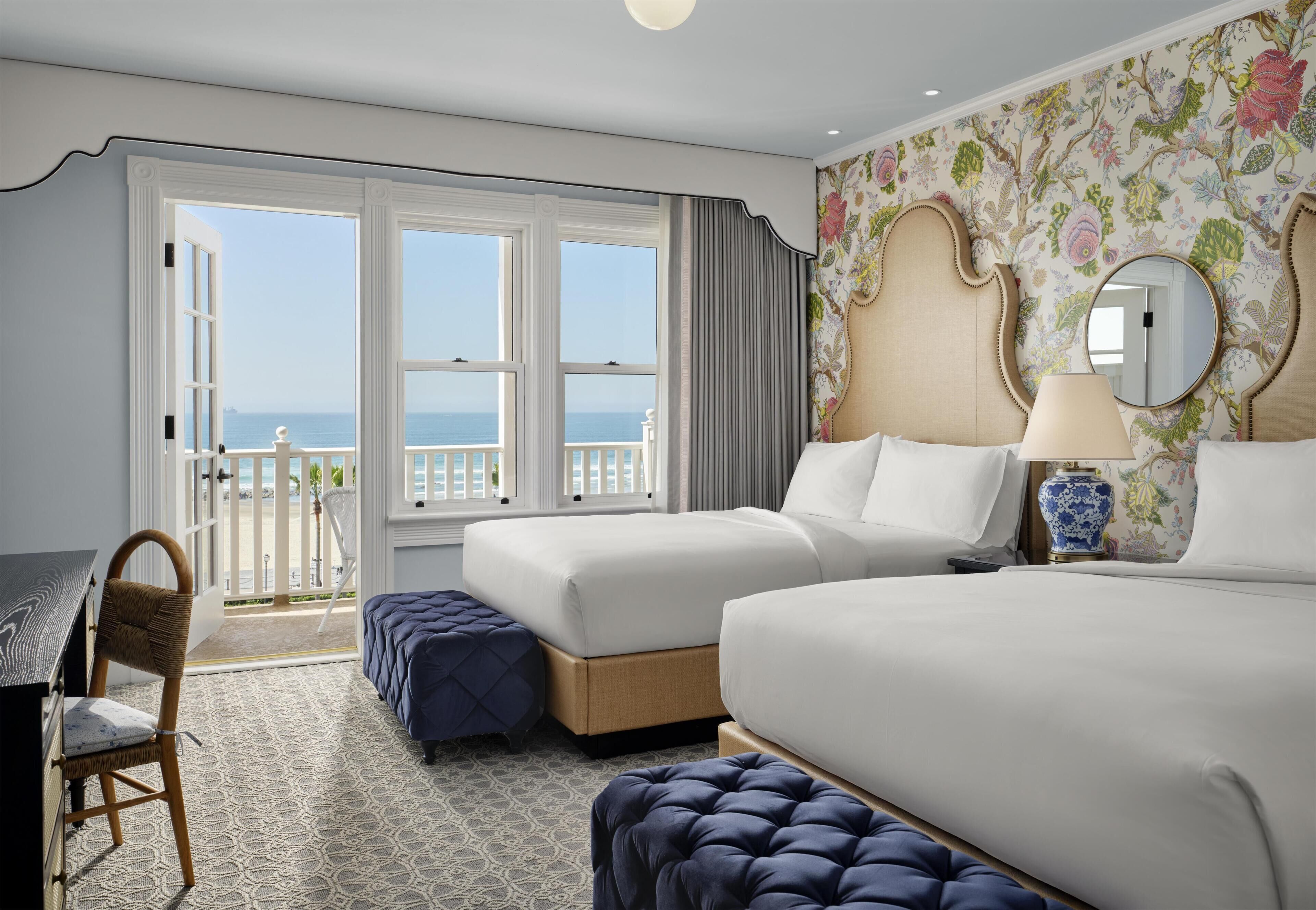 A hotel room with two beds, a desk, a chair, and a balcony with a view of the sea.