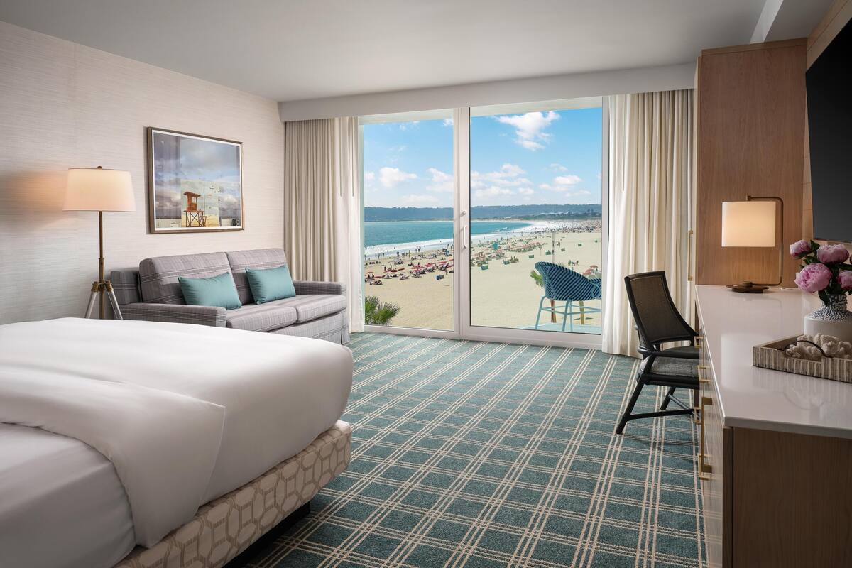 Une chambre d’hôtel avec un lit, un bureau, une chaise, un canapé, une grande fenêtre donnant sur la plage et un tableau au mur.