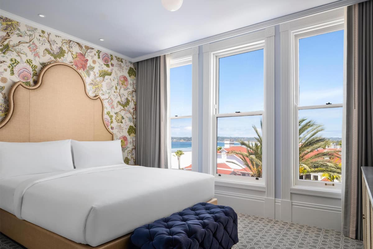 Une chambre d’hôtel avec un grand lit, un pouf capitonné, un papier peint à motifs floraux et une vue sur la plage.