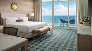 A hotel room with a bed, a desk, a chair, a bench, a large window with a view of the sea, and a balcony with chairs.