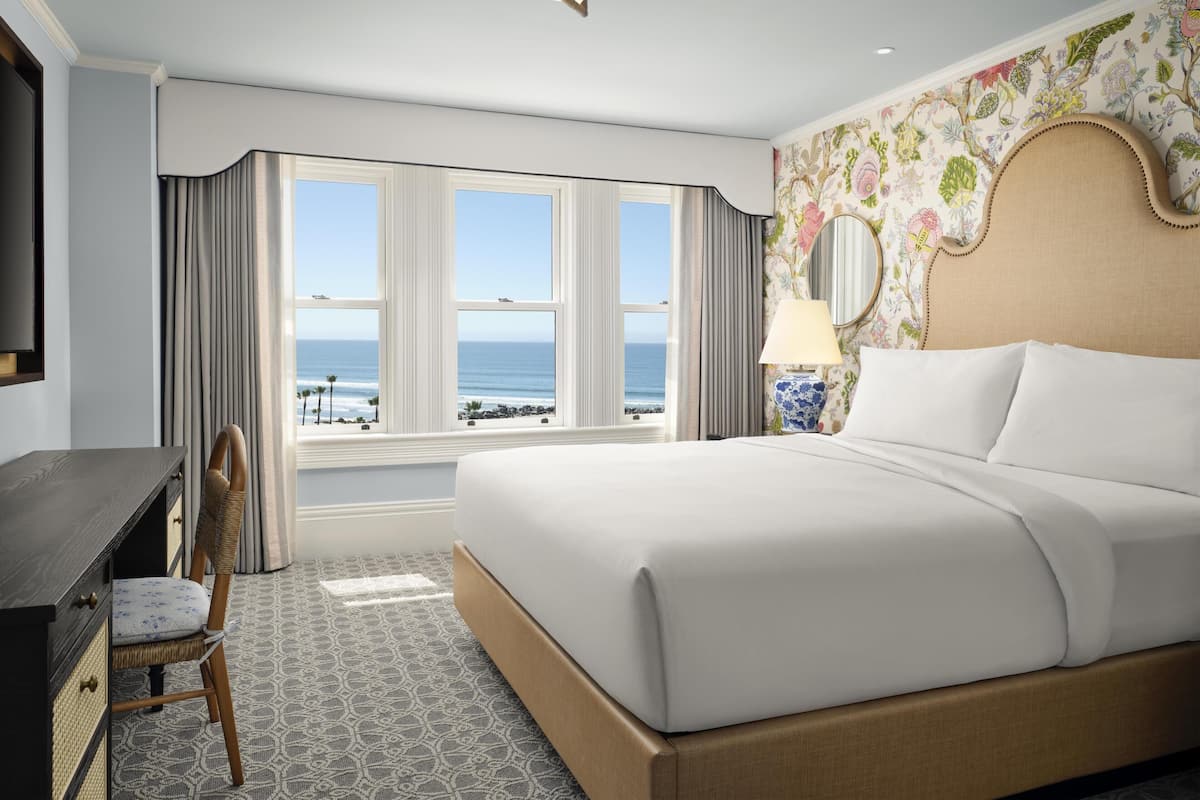 Une chambre d’hôtel avec un grand lit, un bureau, une chaise, une fenêtre donnant sur l’océan et un papier peint à motifs floraux.