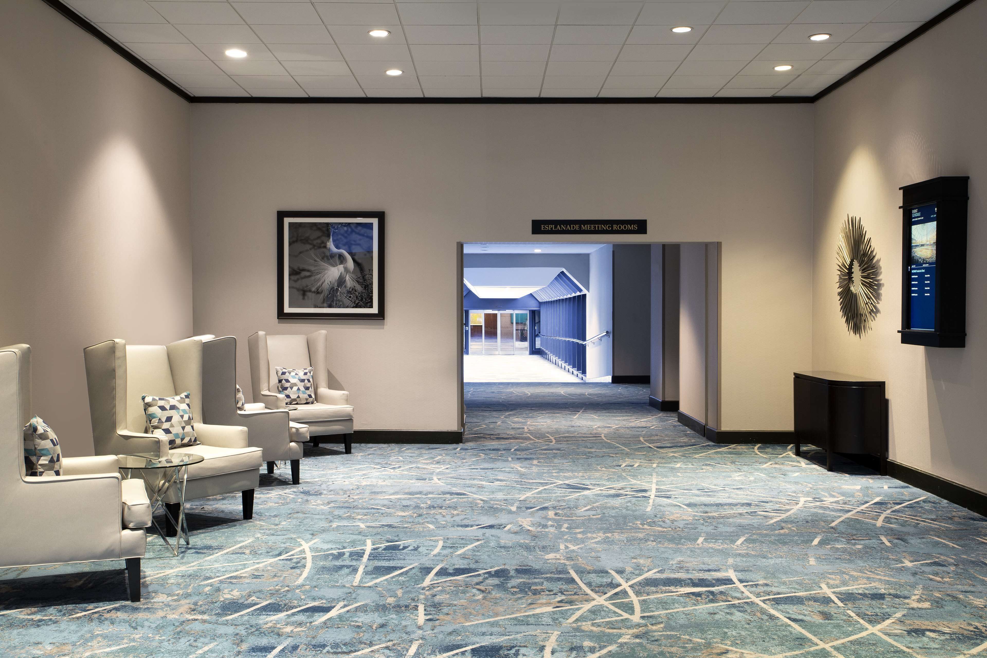 Un hall d’hôtel avec un tapis bleu, un espace détente avec des fauteuils, un tableau encadré au mur et un panneau indiquant l’emplacement des salles de réunion.