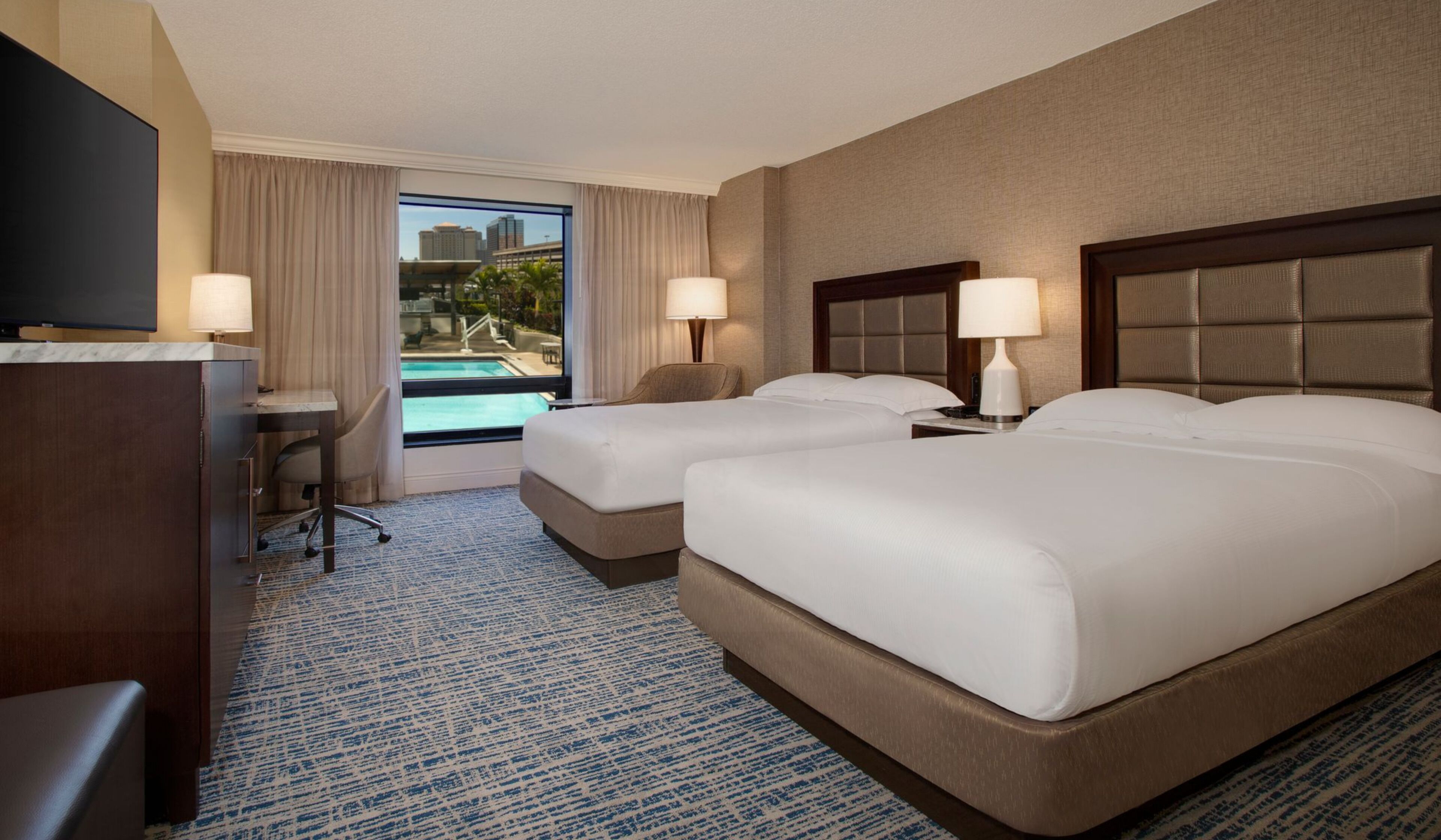 A hotel room with two beds, a desk, a TV, and a view of a swimming pool.