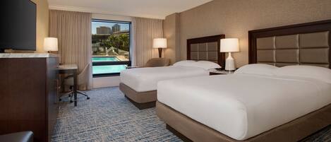 A hotel room with two beds, a desk, a TV, and a view of a swimming pool.