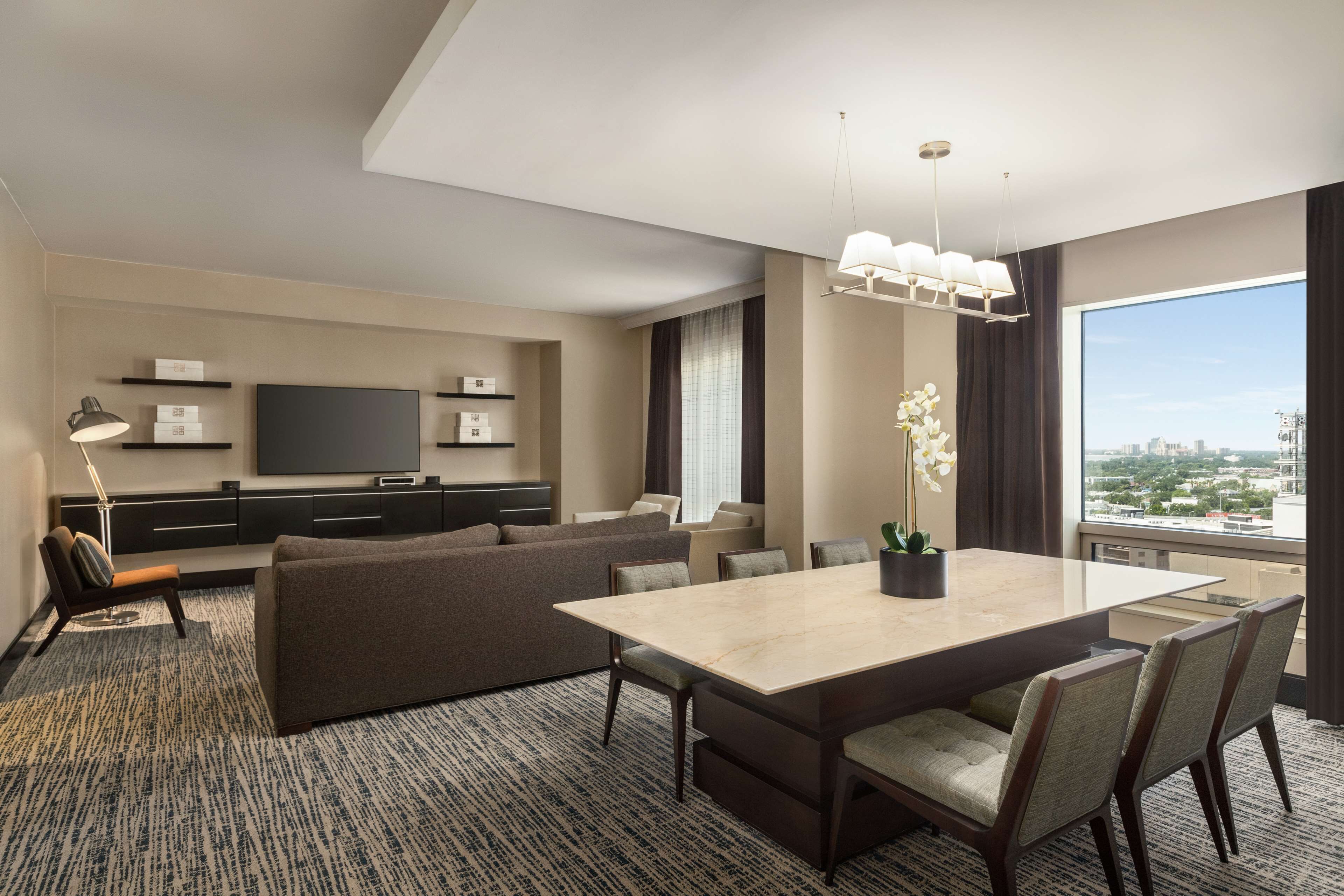 Una habitación de hotel moderna con un amplio comedor, asientos cómodos, un televisor de pantalla plana y vistas a la ciudad.