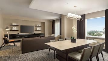 A modern hotel room with a large dining table, comfortable seating, a flat-screen TV, and a city view.