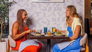 Duas pessoas sentadas à mesa com pratos de comida e bebidas, em um quarto com papel de parede estampado e uma planta ao fundo.