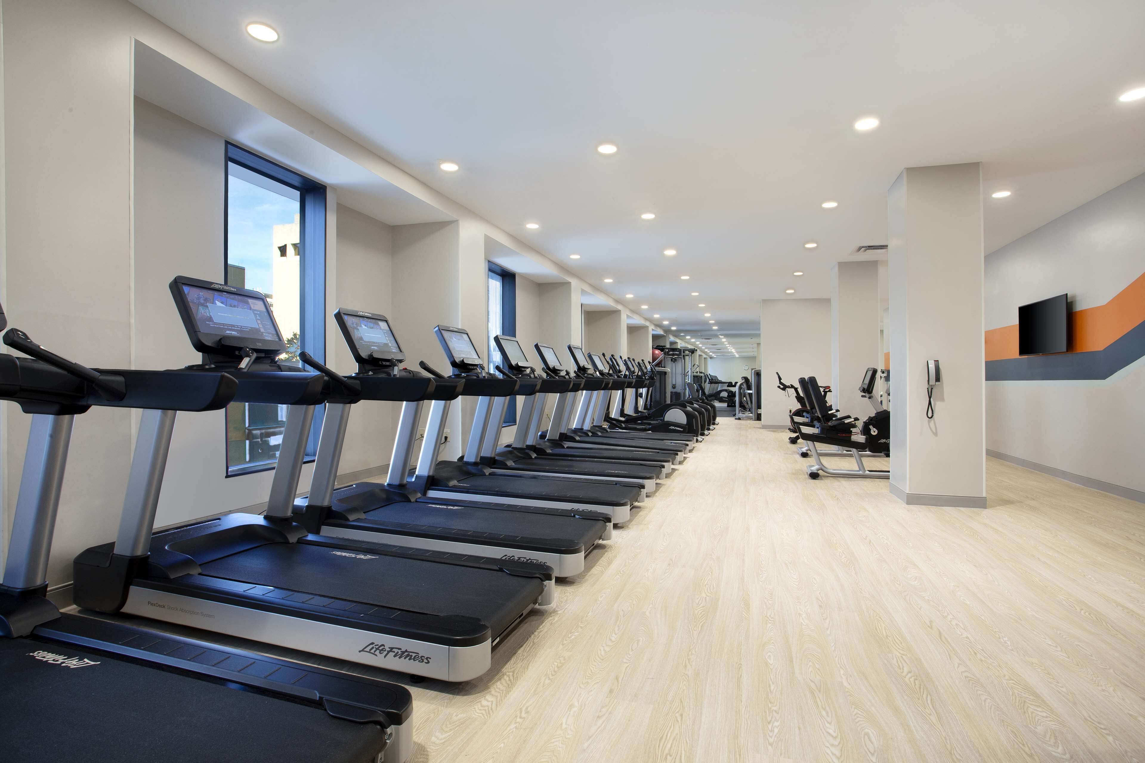 A gym with multiple treadmills lined up against the wall, a wooden floor, and a TV mounted on the wall.