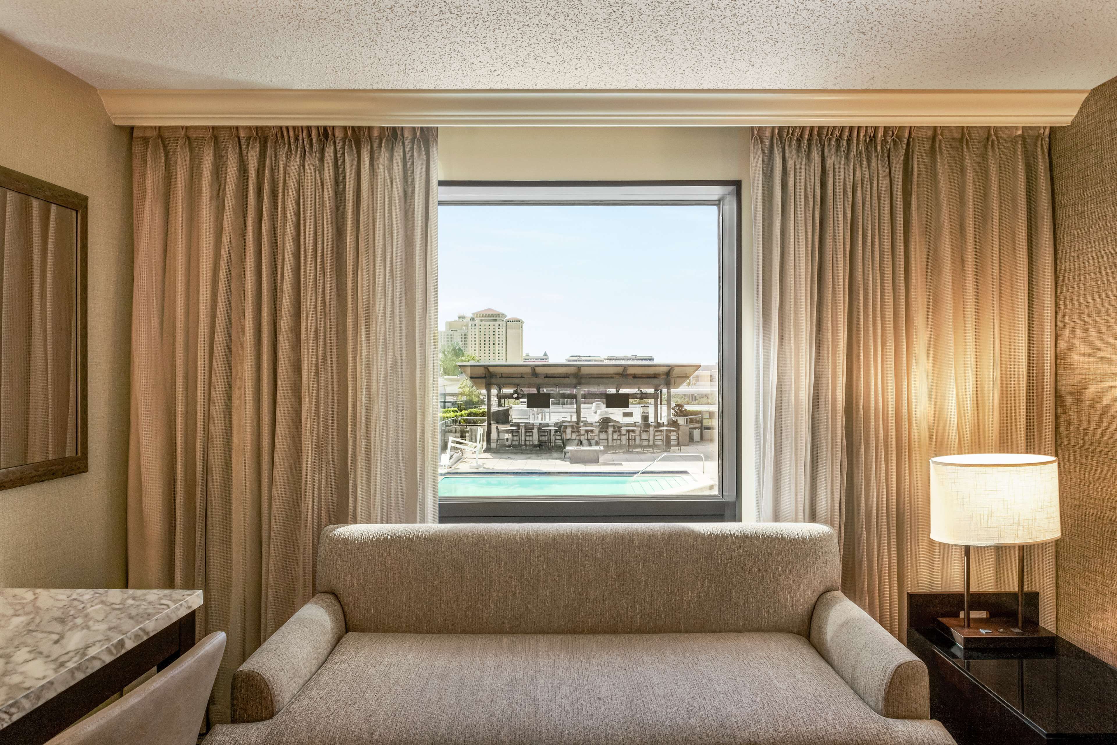 A hotel room with a large window, a sofa, a table, and a lamp.
