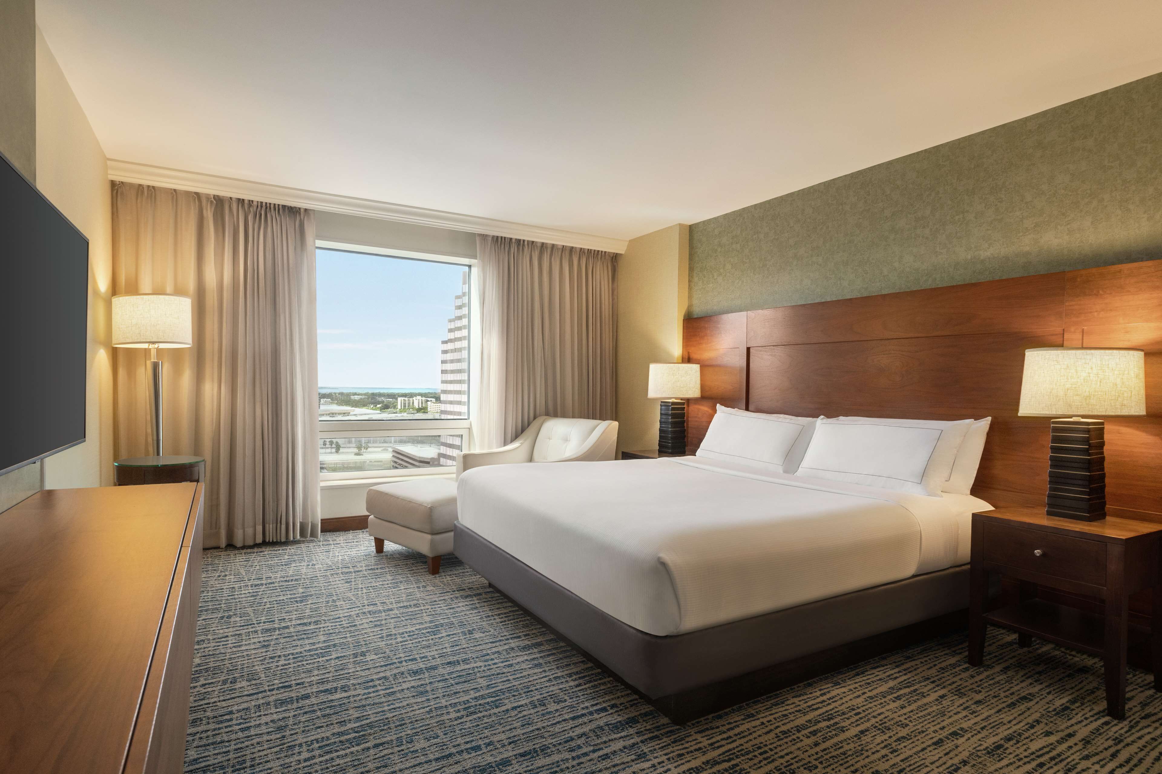 A hotel room with a large bed, two bedside tables with lamps, a chair, a desk, and a view of the city.