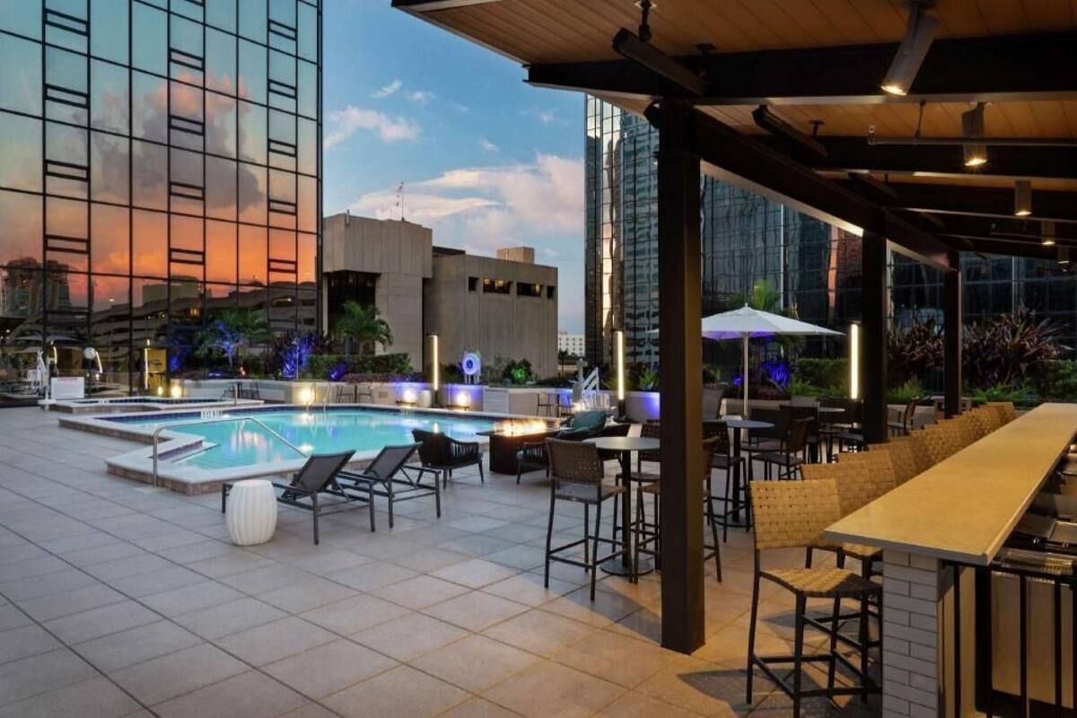 A rooftop pool area with lounge chairs, a bar, and a view of the city skyline.
