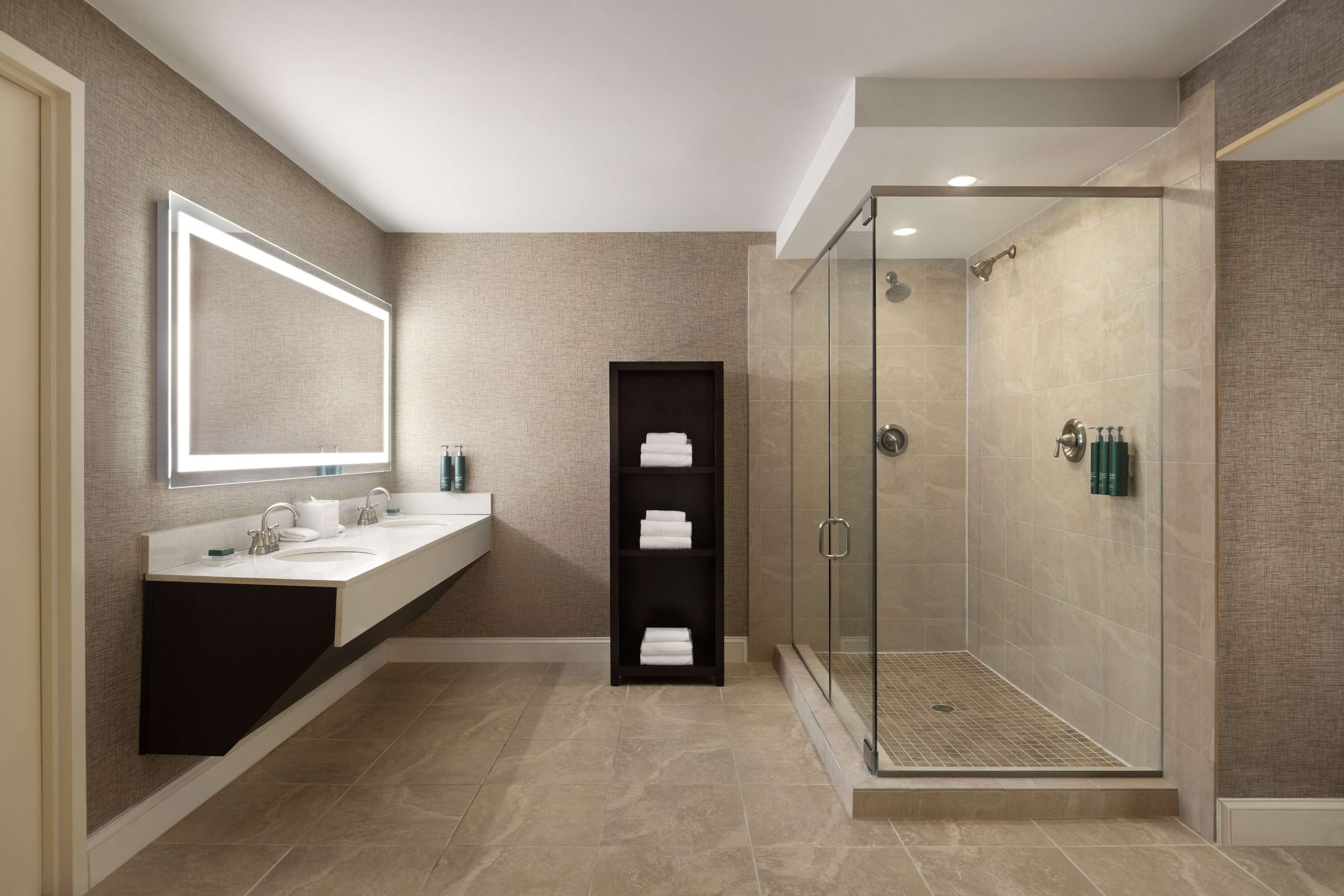 A modern bathroom with a large mirror, two sinks, a glass-enclosed shower, and a shelf with towels.