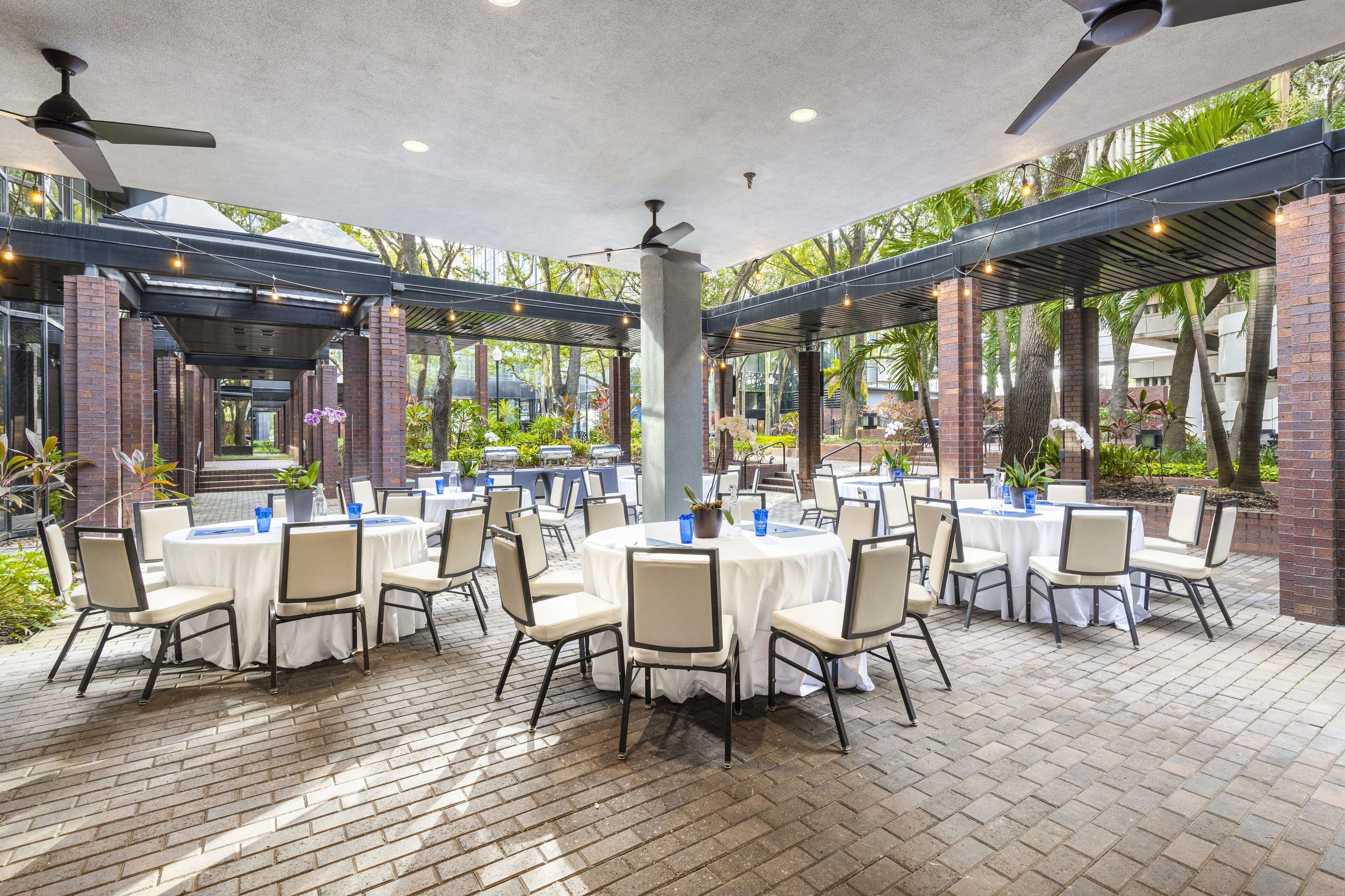 A covered outdoor dining area with multiple round tables and chairs, surrounded by brick pillars and greenery.