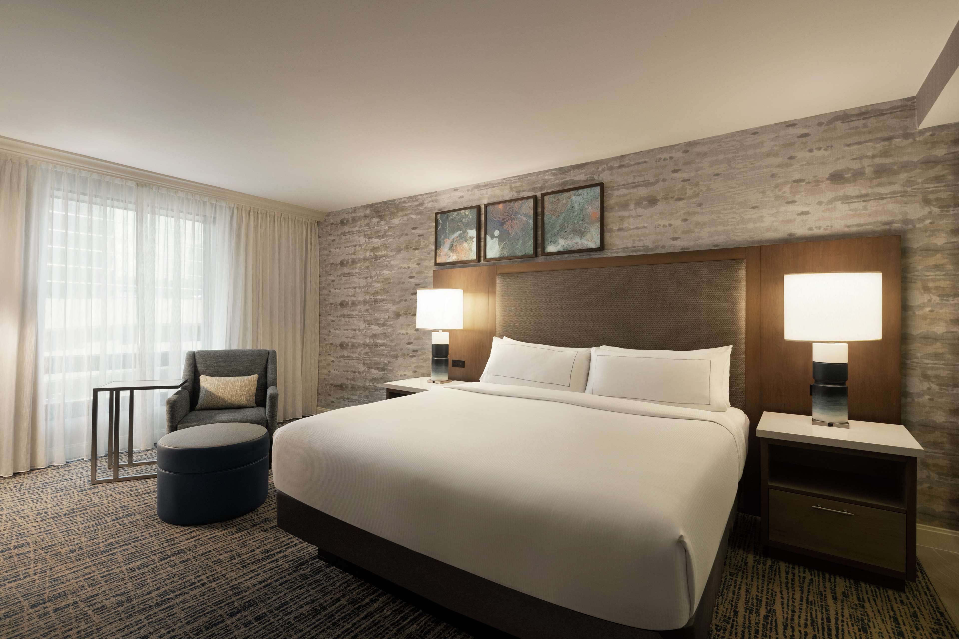 A large bed with white linens, two bedside tables with lamps, a dark armchair, and a round ottoman.