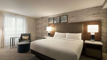 A large bed with white linens, two bedside tables with lamps, a dark armchair, and a round ottoman.
