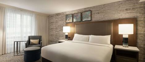 A large bed with white linens, two bedside tables with lamps, a dark armchair, and a round ottoman.