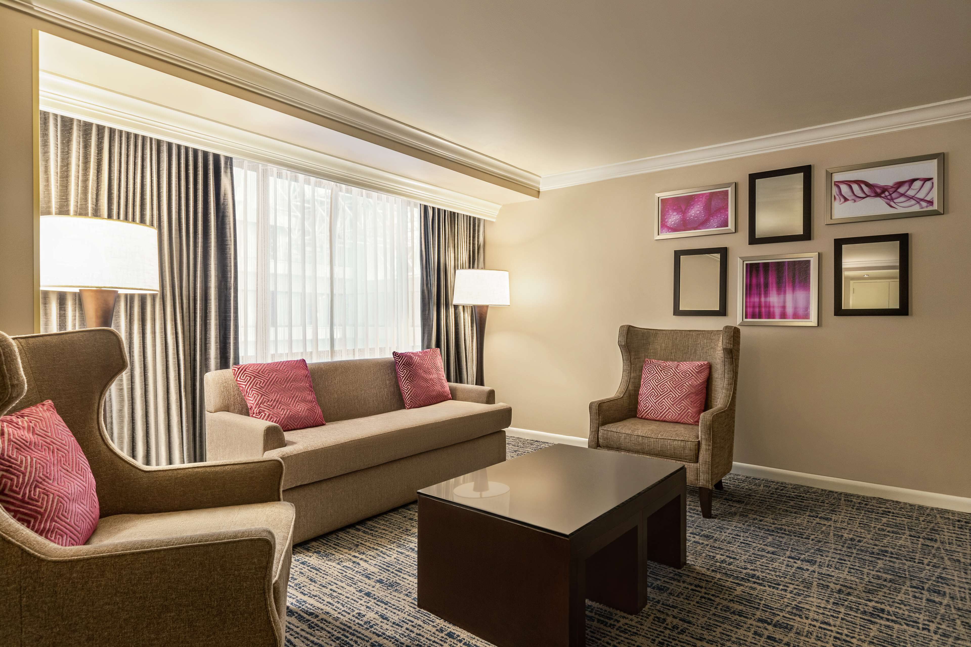 A hotel room with a sofa, armchairs, a coffee table, and a large window with curtains. There are framed pictures on the wall.