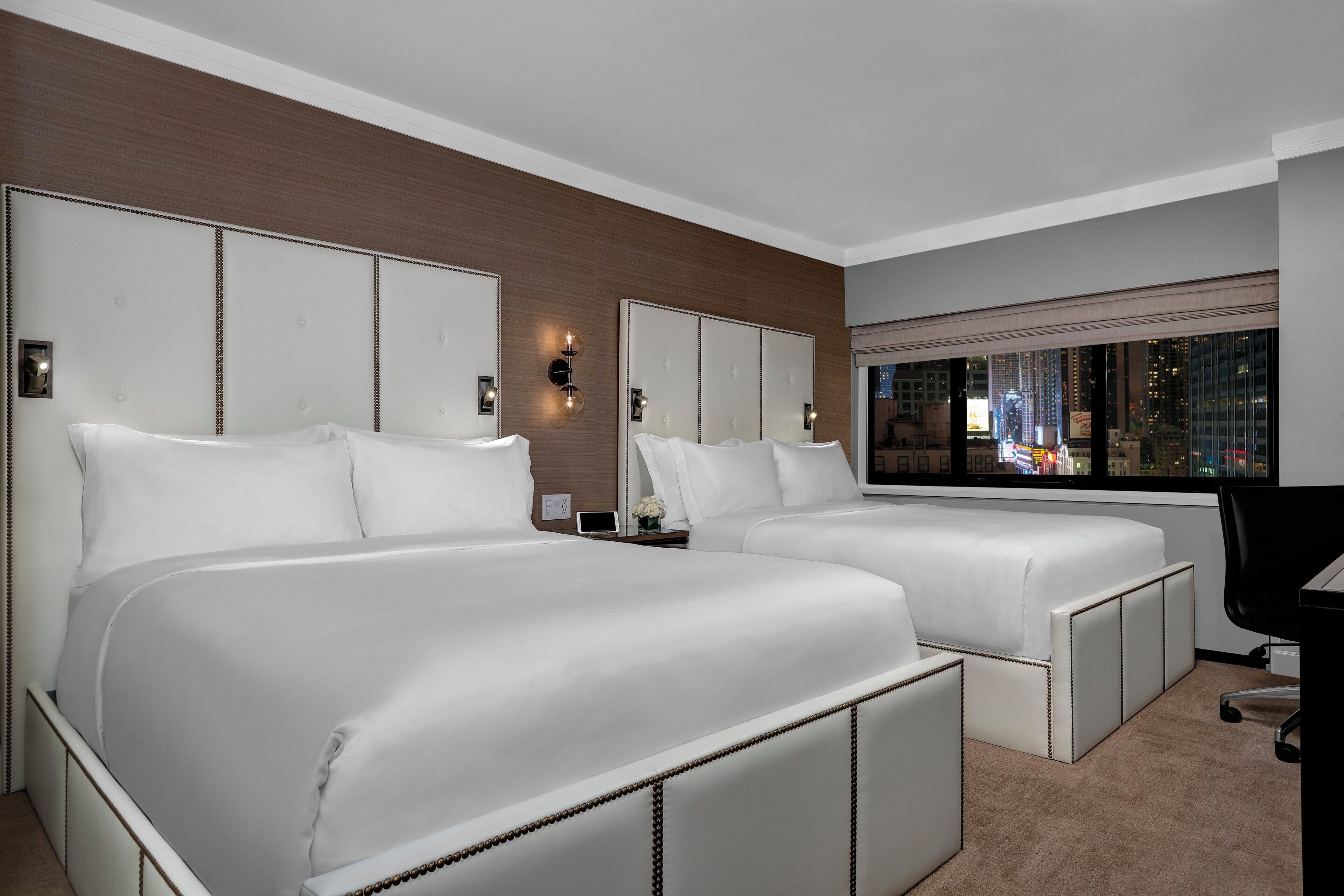 Habitación de hotel con dos camas, un ventanal grande con vista a la ciudad y un escritorio con una silla.