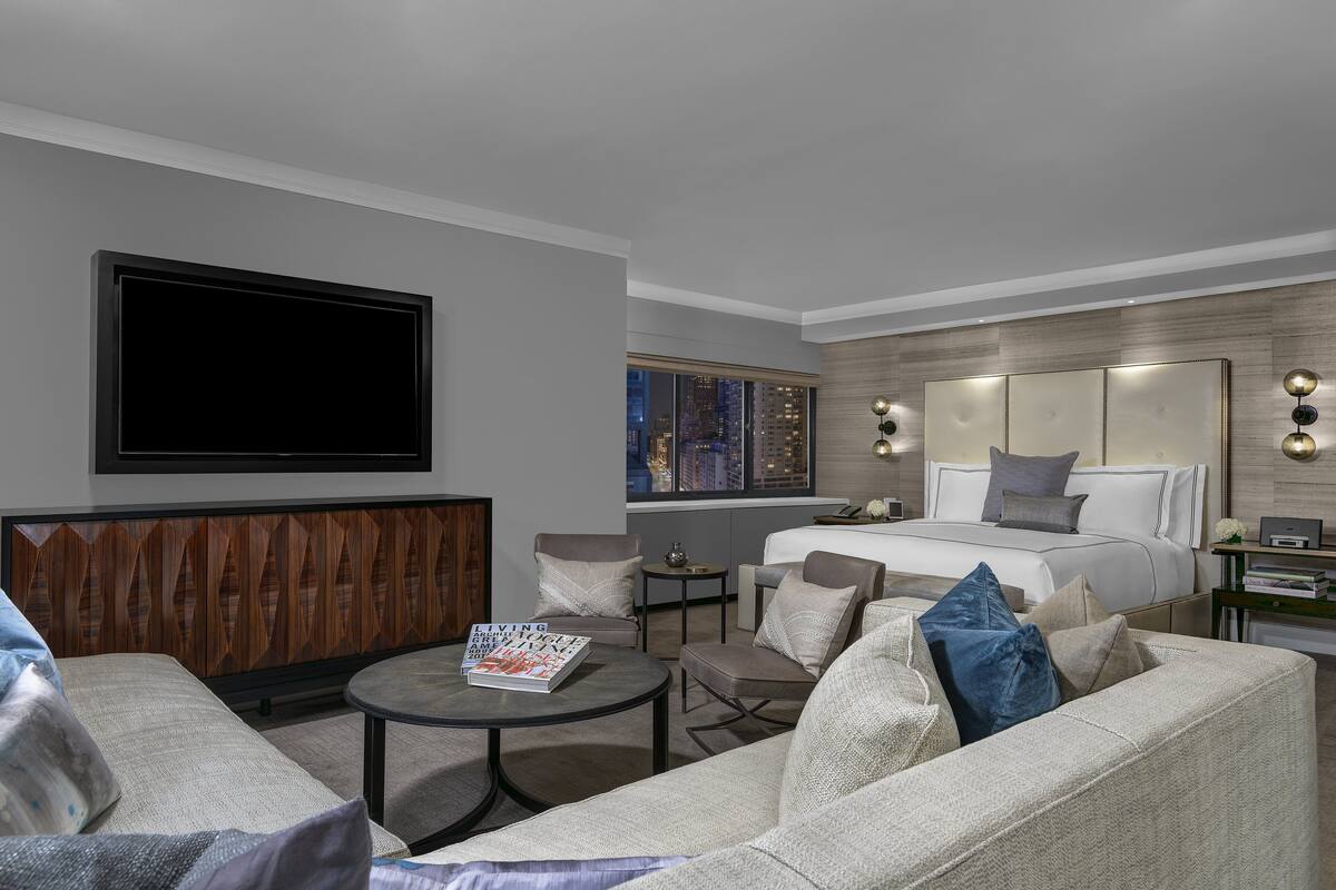 A modern hotel room with a large bed, a seating area with a coffee table, a television, and a city view.