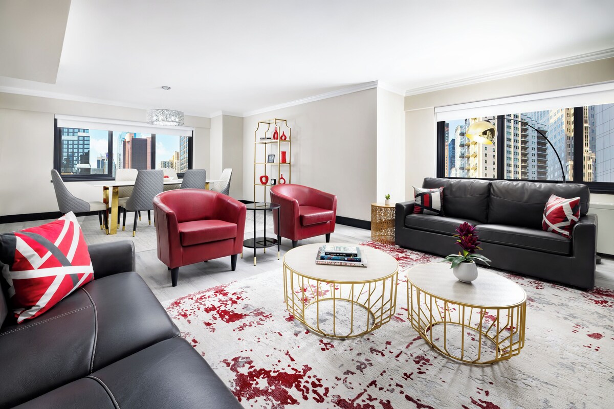 Een moderne woonkamer met een zwarte leren bank, rode fauteuils, een eethoek met een witte tafel en een stadszicht door grote ramen.
