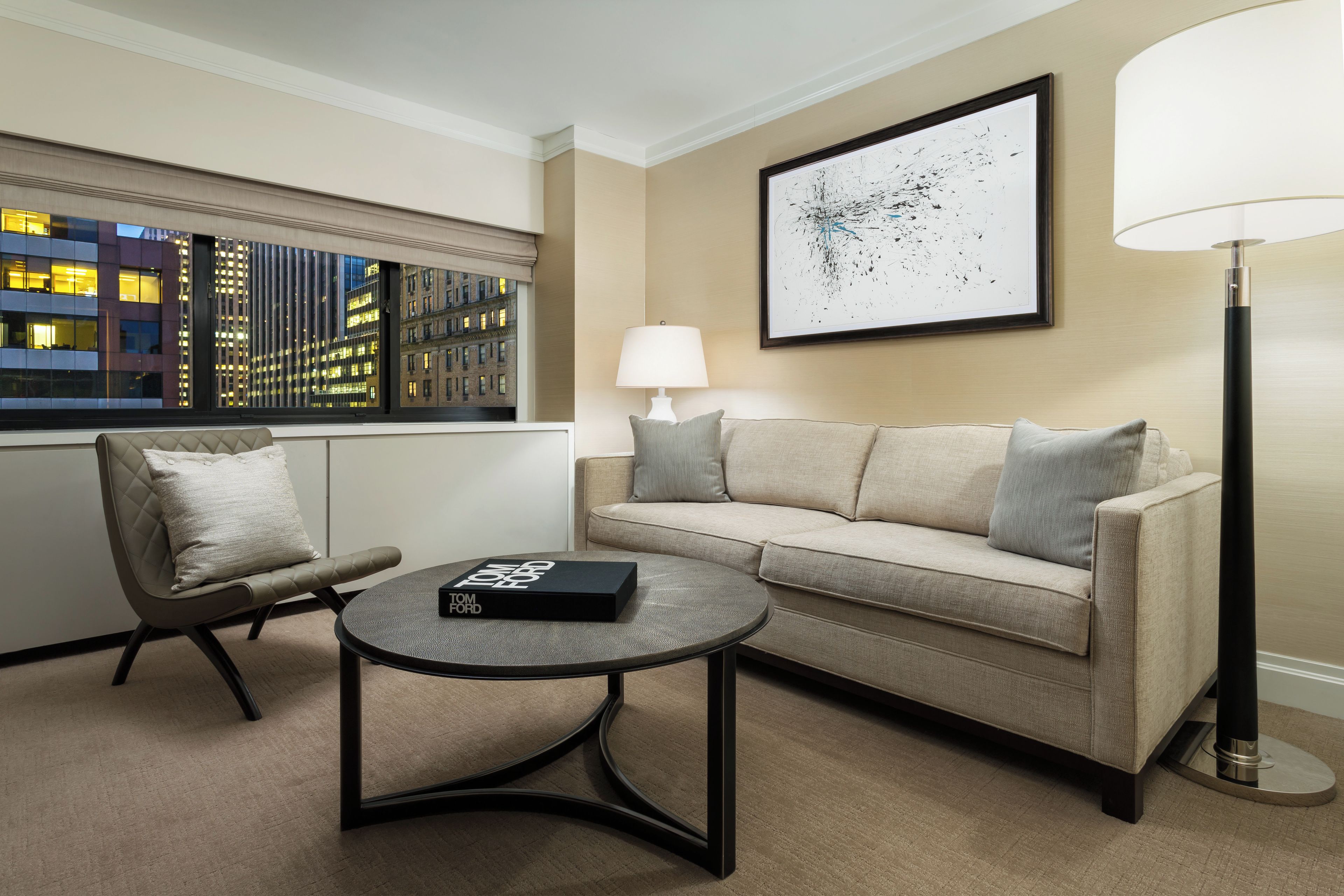 A hotel room with a sofa, armchair, round coffee table, and a lamp.