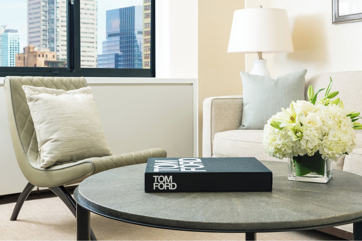 A modern living room with a round coffee table, a book titled 'TOM FORD', a vase of white flowers, and a city view through a window.