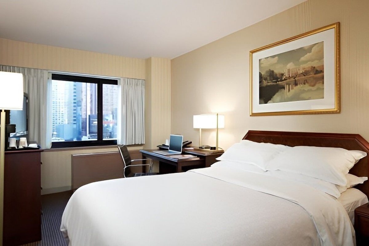 A hotel room with a large bed, a desk with a laptop, a chair, a desk lamp, a nightstand, and a window with curtains.