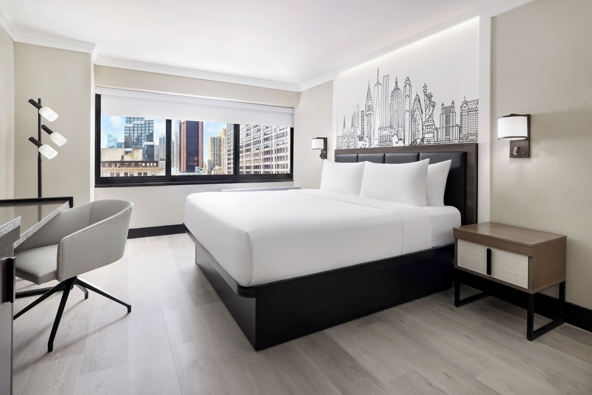 A modern hotel room with a large bed, a desk, and a cityscape view.