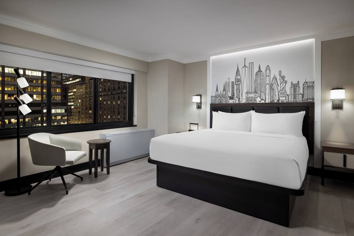 A modern hotel room with a large bed, a city skyline mural, a city view through the window, a small table, and a chair.