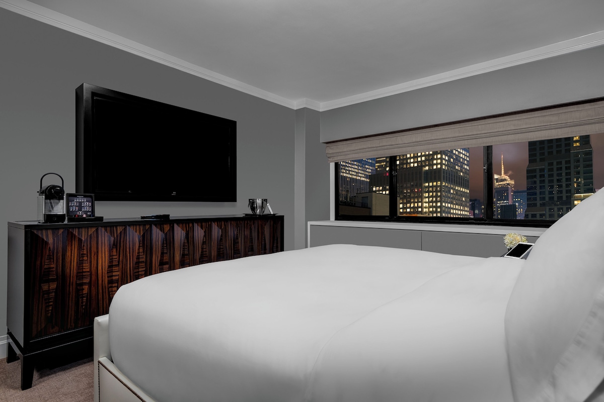 Een hotelkamer met een groot bed, een flatscreen televisie, een houten dressoir en een raam met uitzicht op de stad.
