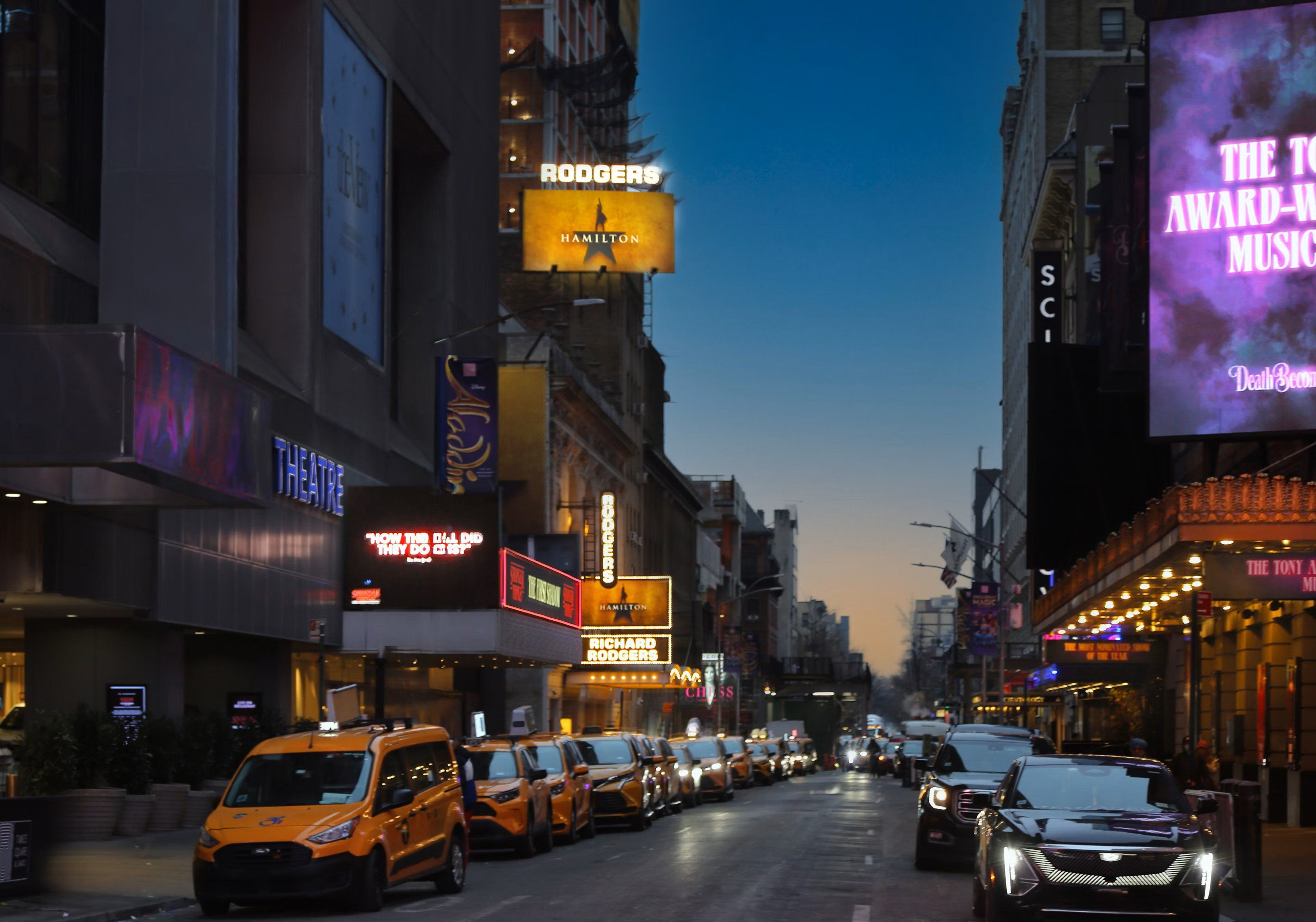 Une rue bordée de théâtres illuminés et de panneaux publicitaires, dont des enseignes pour les spectacles « Rodgers » et « Hamilton ».