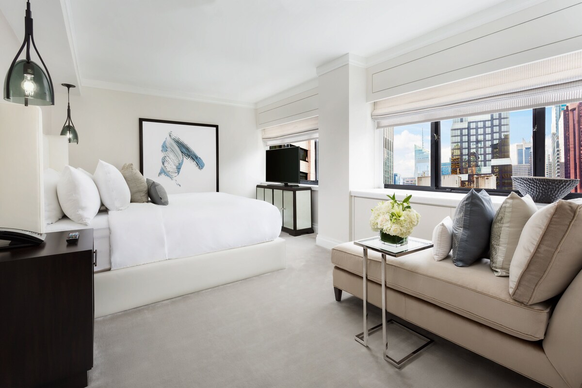 Una habitación de hotel moderna con una cama grande, un sofá, una mesita con un jarrón de flores y vistas a la ciudad por la ventana.
