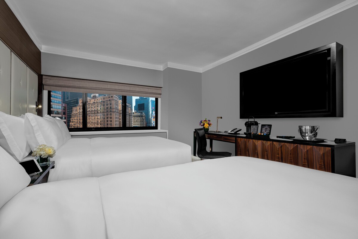 Een hotelkamer met een groot bed, een bureau met een flatscreen televisie, een raam met uitzicht op de stad en een vaas met bloemen.