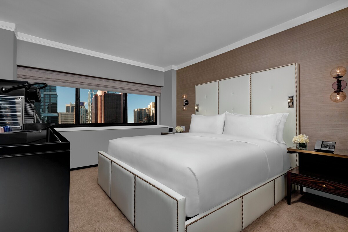 A modern hotel room with a large bed, a desk, and a view of the city skyline.