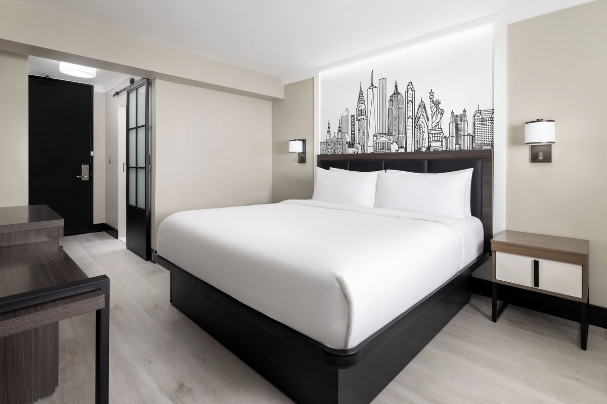 A modern hotel room with a large bed, a desk, and a cityscape wall art.
