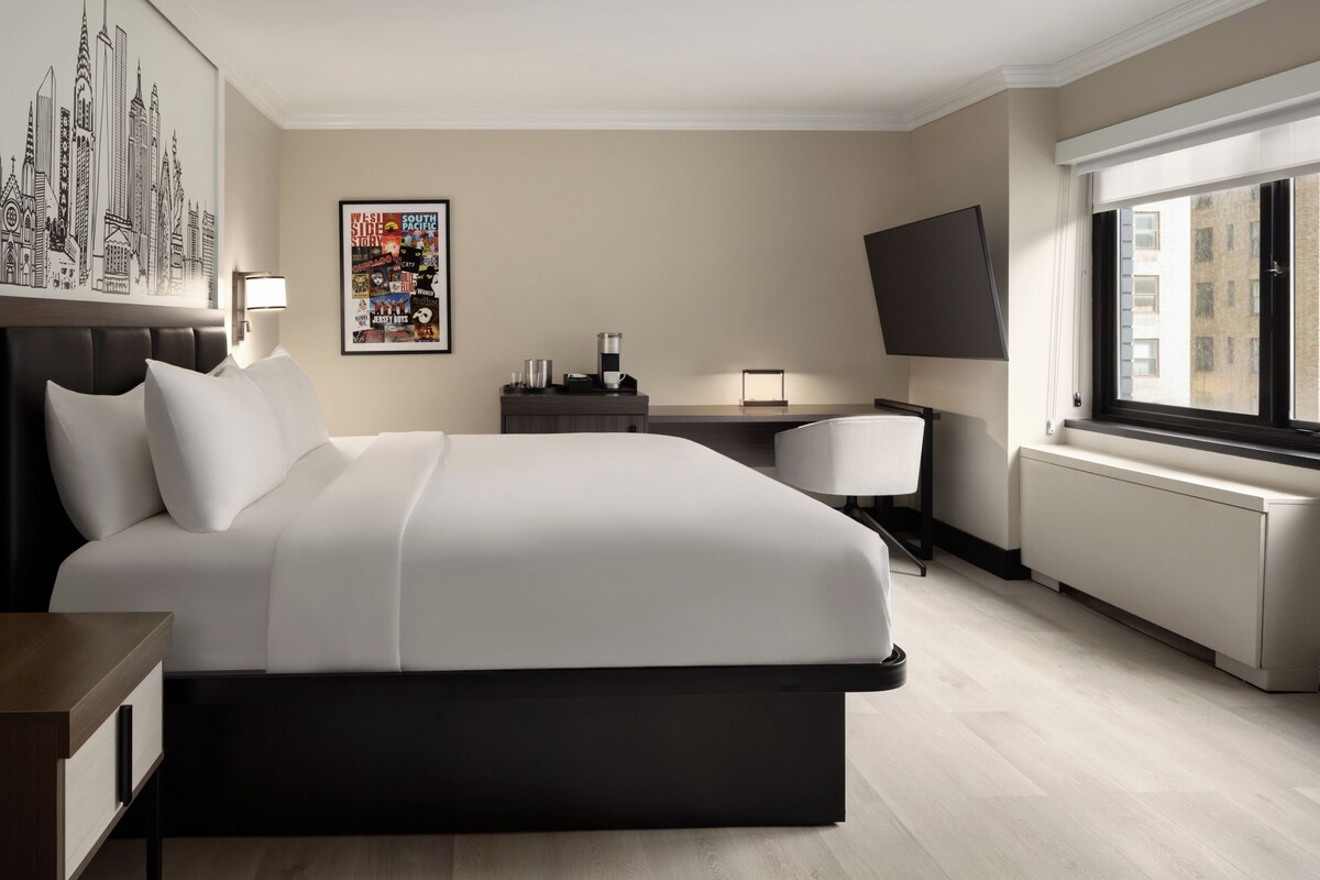 Quarto de hotel moderno com uma cama grande, uma escrivaninha, uma cadeira, uma TV de tela plana e um pôster na parede.