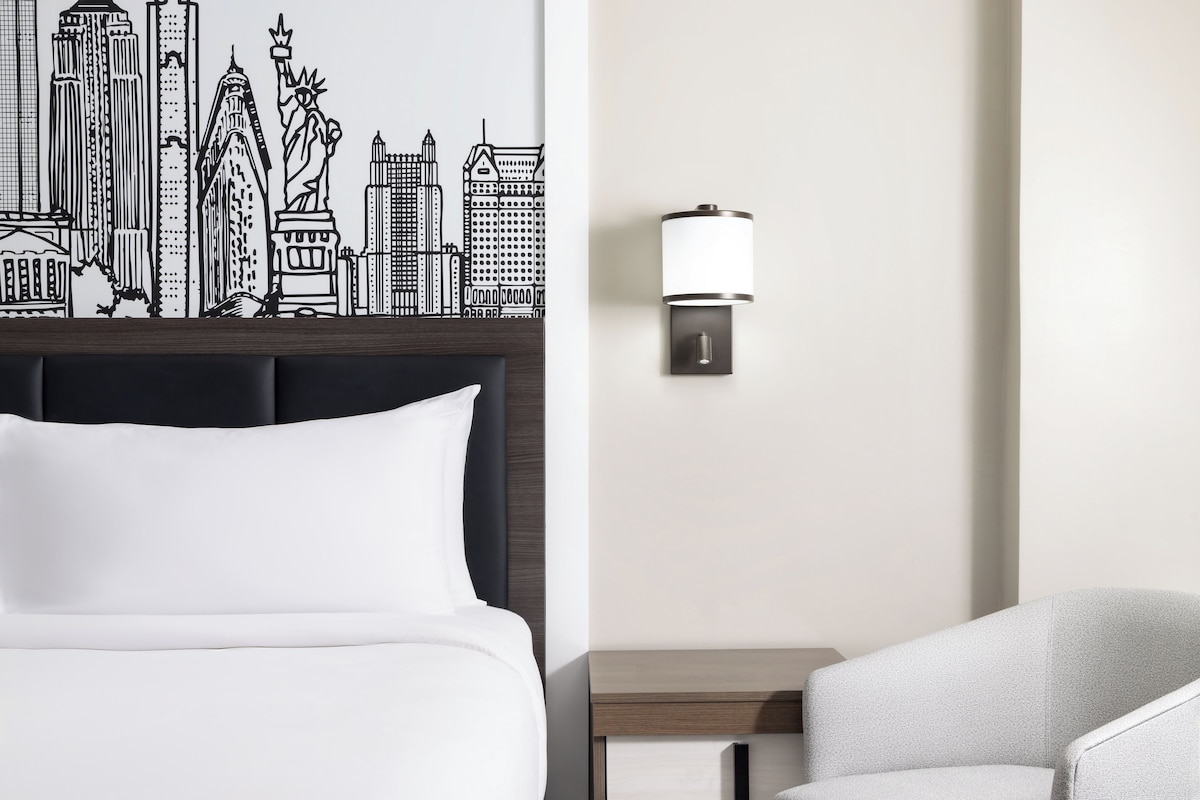 A hotel room with a bed, a nightstand, a lamp, and a wall with a cityscape mural.