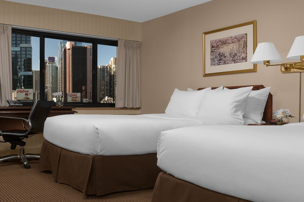 A hotel room with two beds, a desk, a chair, and a large window offering a city view.