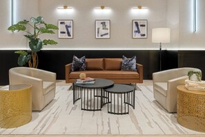 A modern living room with a brown sofa, two armchairs, a coffee table, and a floor lamp. There are framed artworks on the wall and a large plant in the corner. - The Manhattan at Times Square (New York)