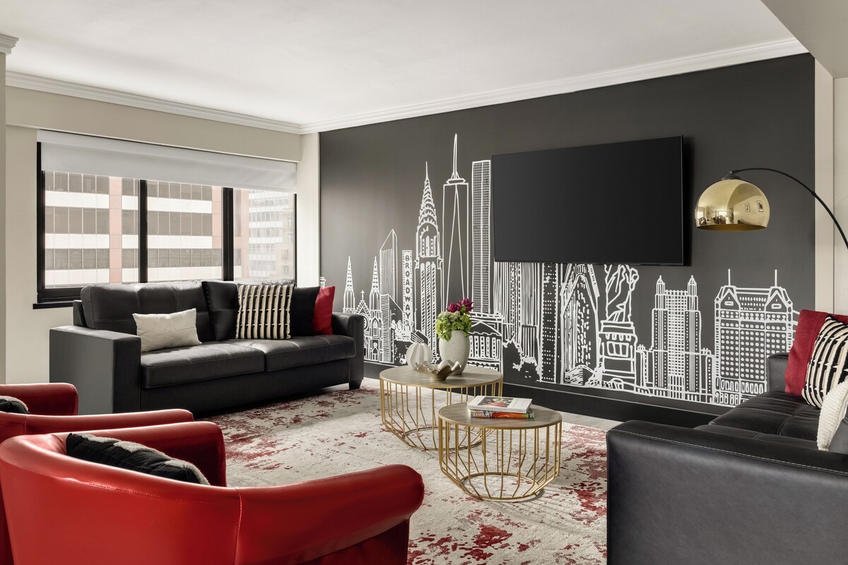 Un salon moderne avec un canapé noir, des fauteuils rouges, une table ronde dorée et une fresque murale représentant un paysage urbain.