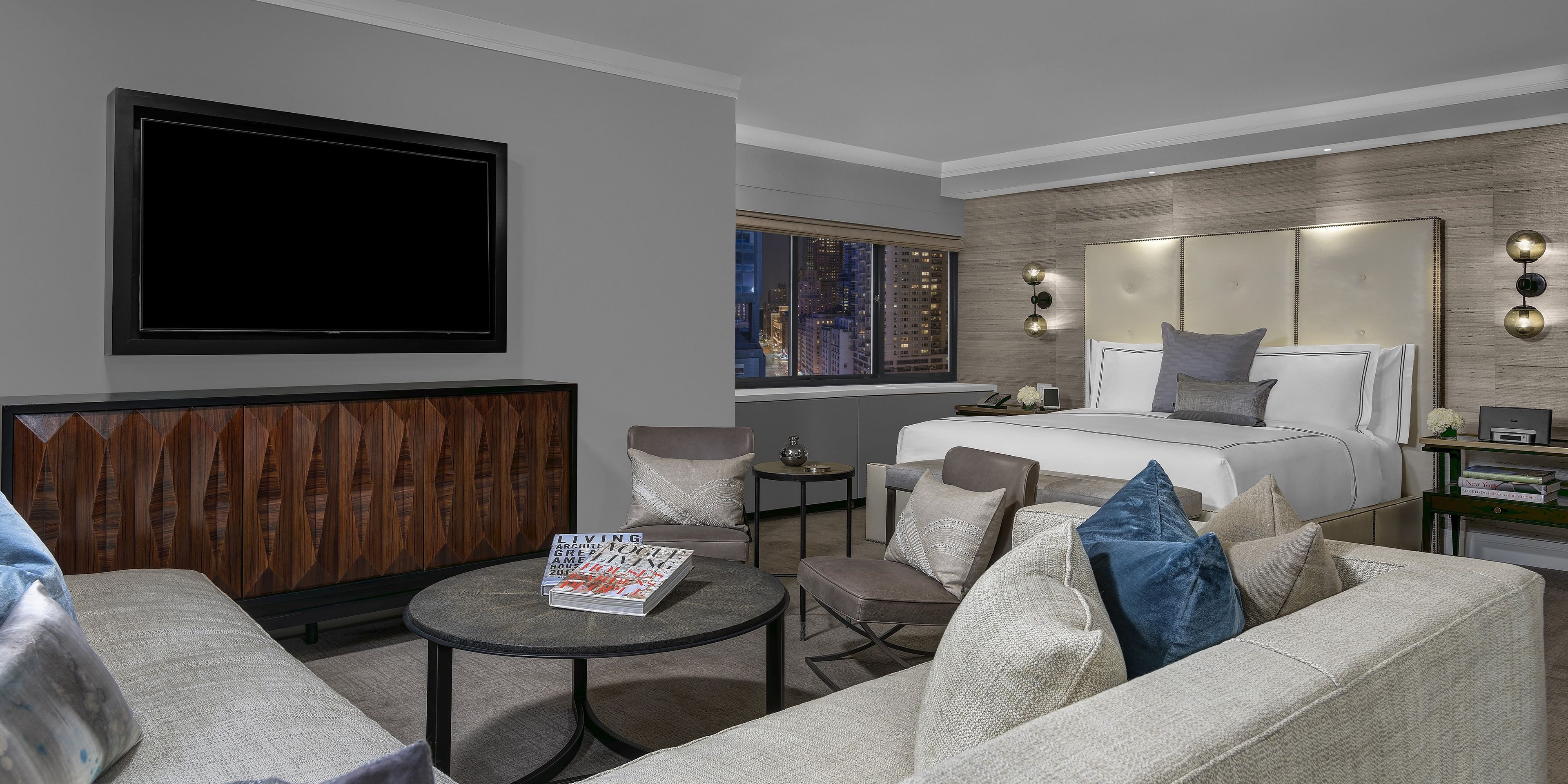 A modern hotel room with a large bed, a seating area with a coffee table, a television, and a city view.
