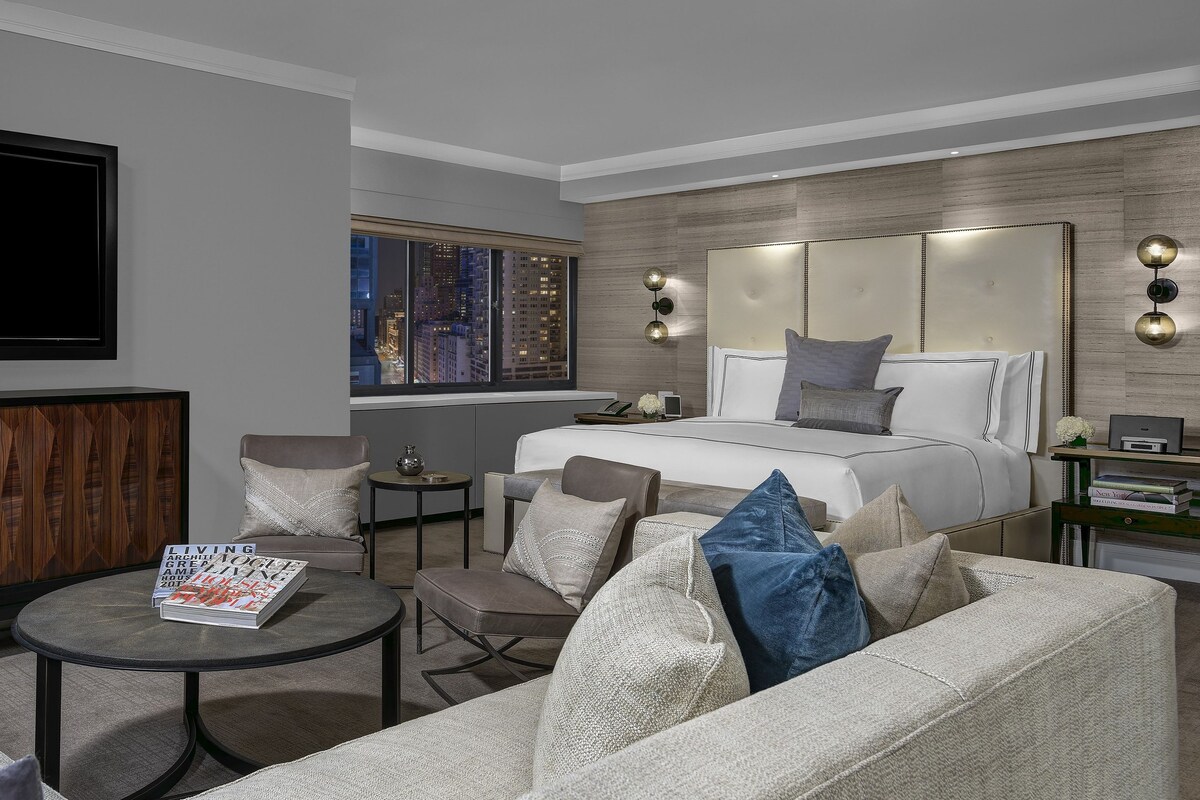 A modern hotel room with a large bed, a seating area with a coffee table, a television, and a city view.