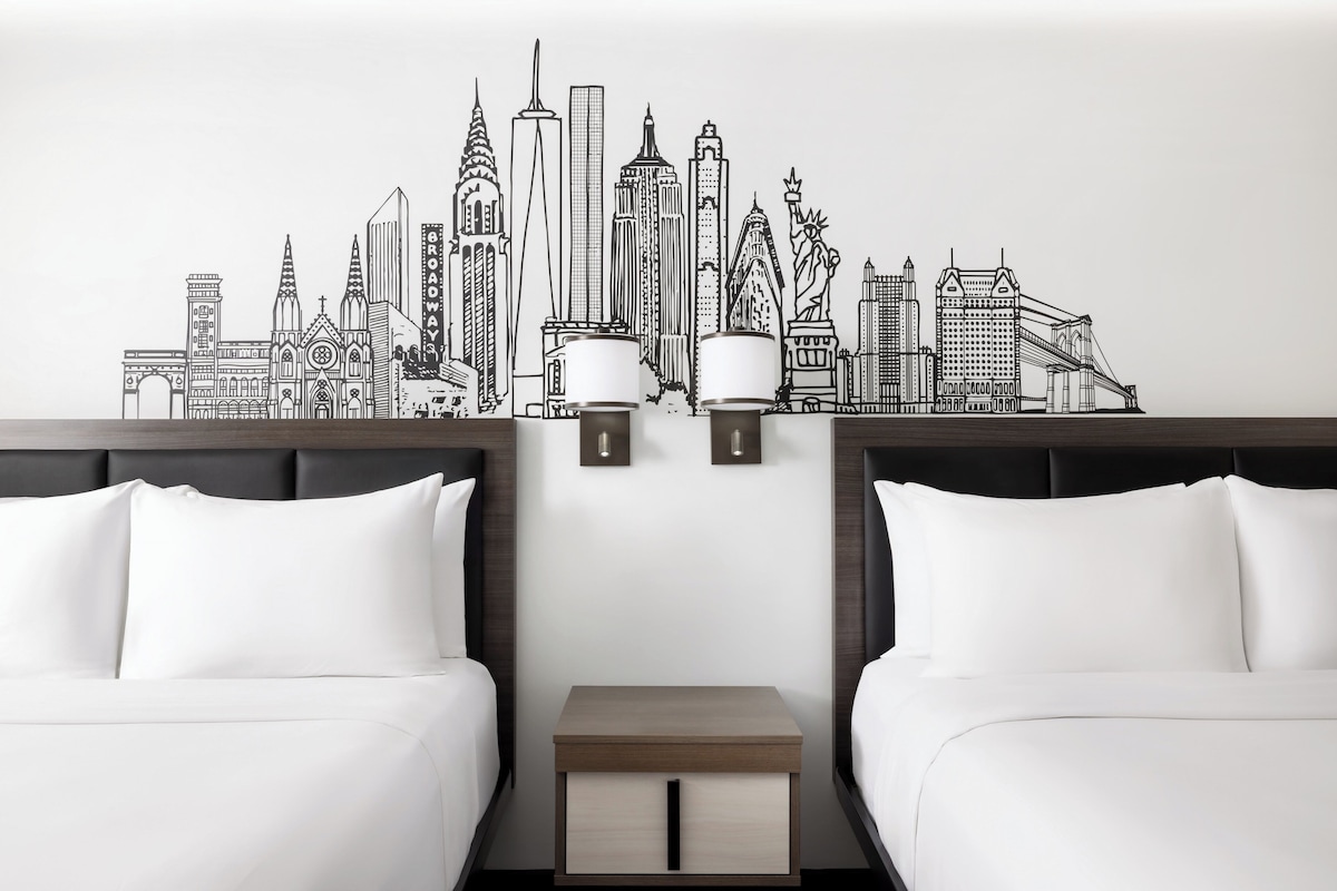 A hotel room with two beds, a nightstand, and a wall mural of a city skyline.