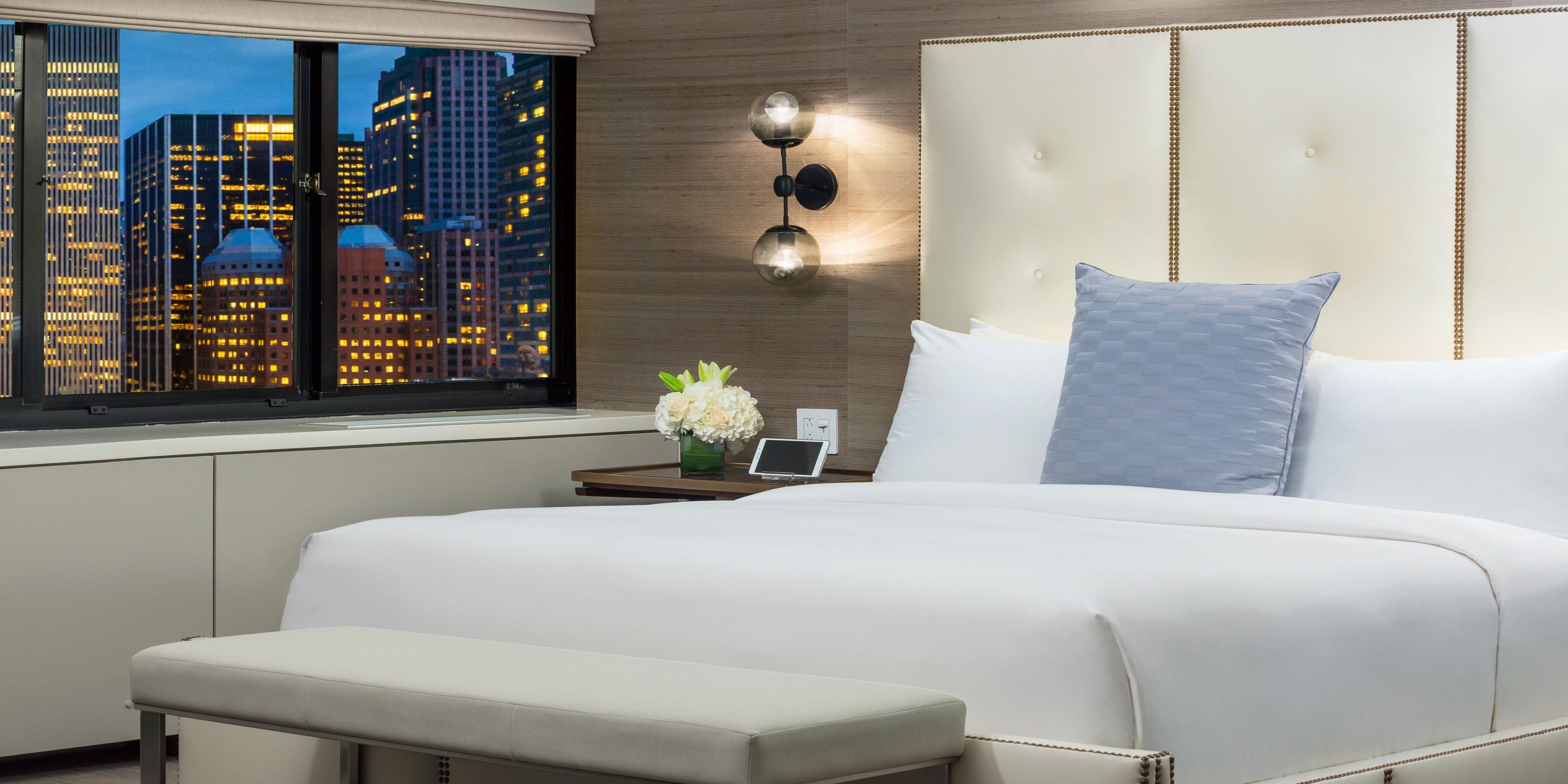 A hotel room with a large bed, a bedside table with a lamp, a small vase of flowers, and a view of city buildings through the window.