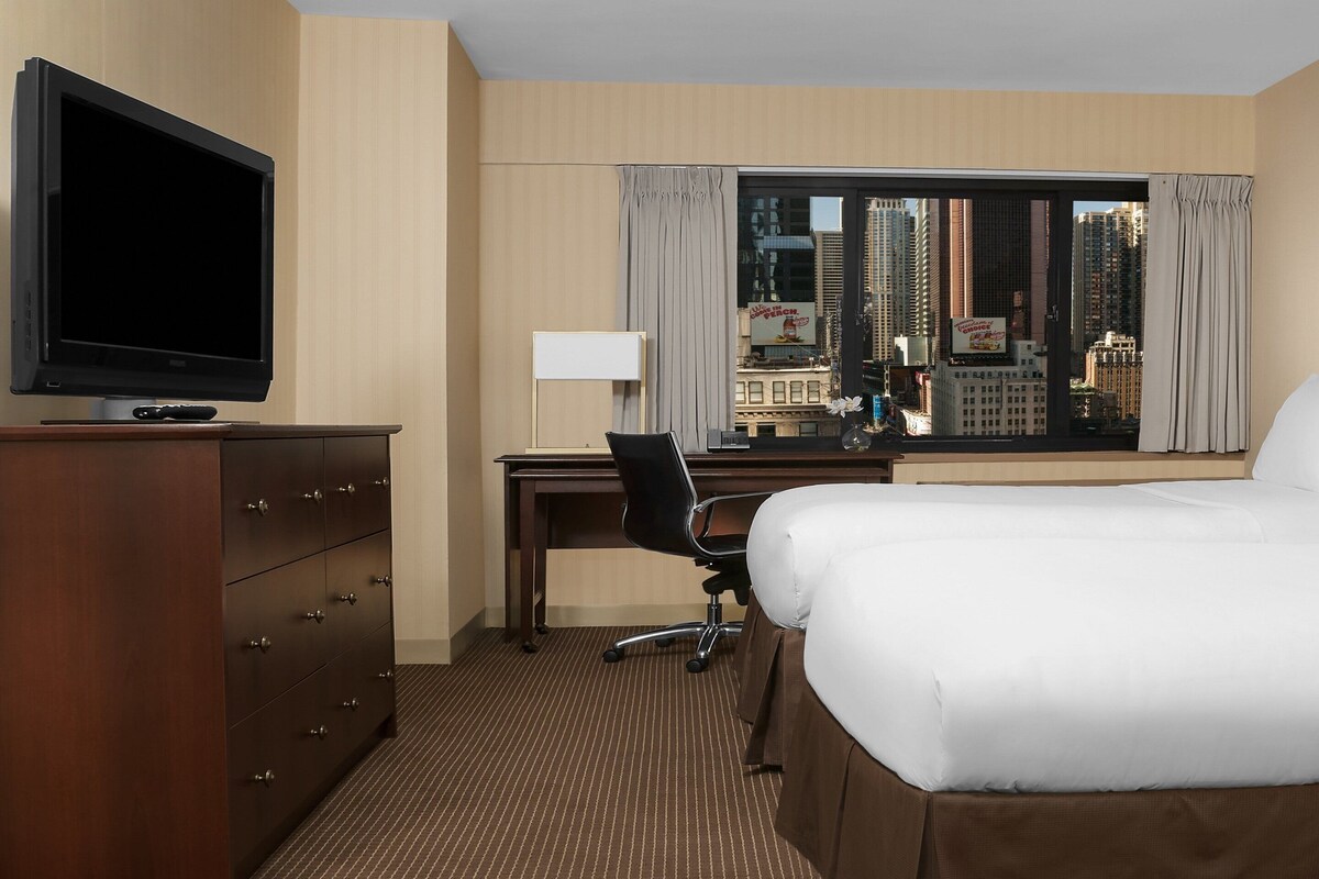 A hotel room with a bed, a desk, a television, and a window with a city view.