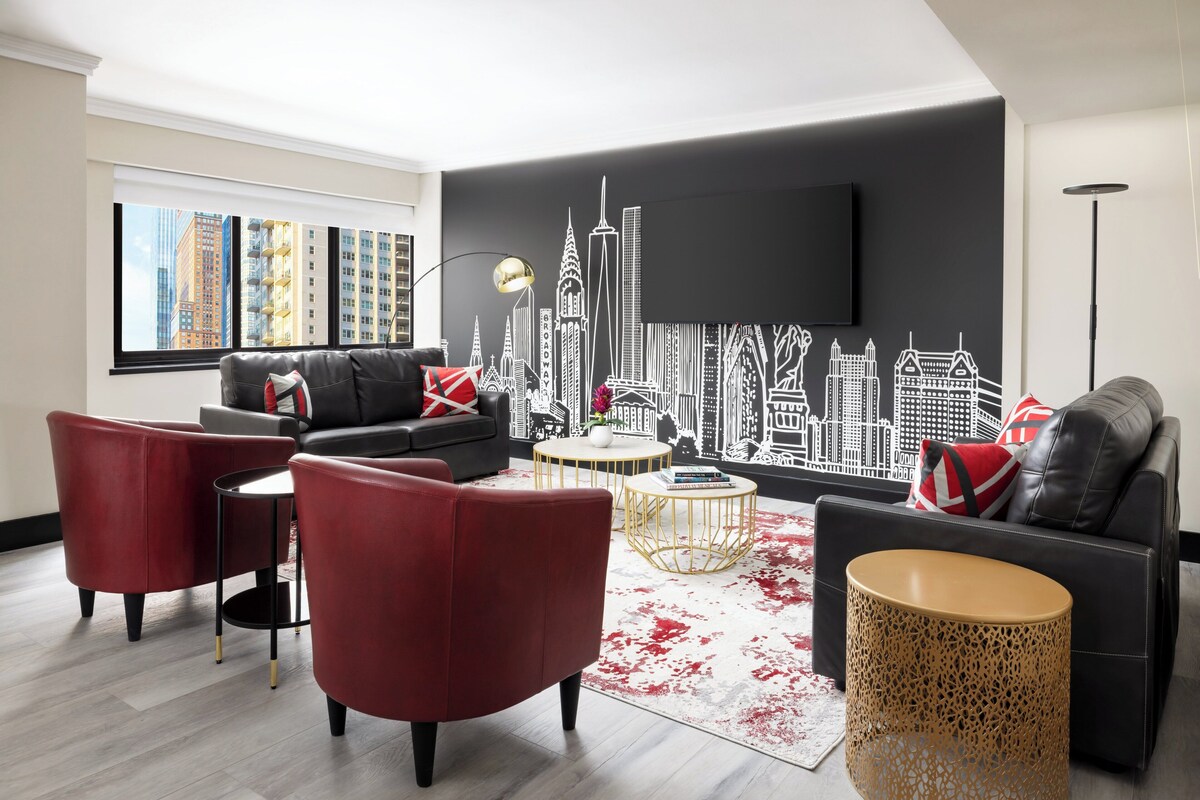 Una sala de estar moderna con un sofá negro, sillas rojas, una mesa de centro redonda y un mural con vistas a la ciudad en la pared.